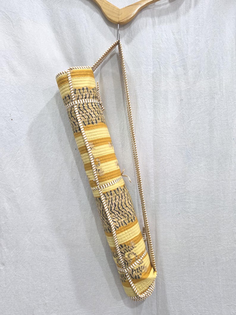 May include: A cylindrical yoga mat bag featuring a quilted texture with alternating horizontal bands of yellow, gold, and white stripes, accented by dark gray leaf-like patterns. The bag includes a long, thin, matching fabric shoulder strap.
