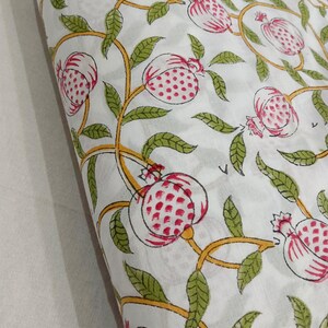 May include: A white fabric featuring a repeating block print pattern of pink pomegranates with red seeds, connected by winding yellow vines and green leaves. The textile displays a consistent botanical motif across the entire visible surface area.