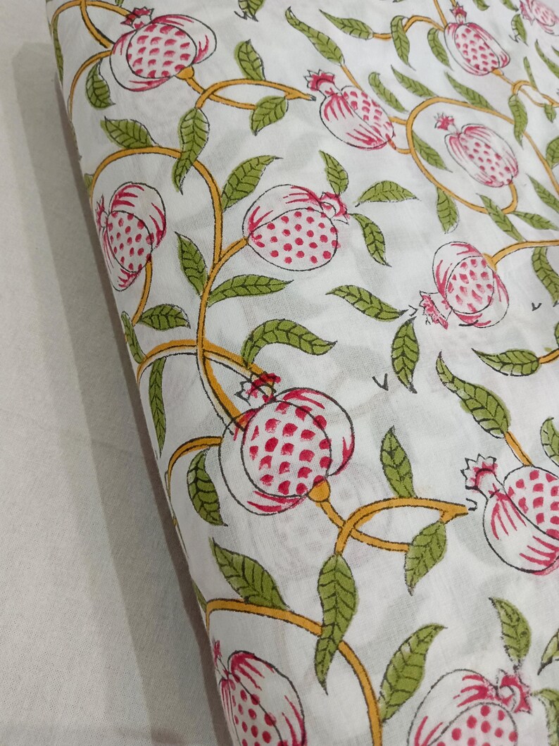 May include: A white fabric featuring a repeating block print pattern of pink pomegranates with red seeds, connected by winding yellow vines and green leaves. The textile displays a consistent botanical motif across the entire visible surface area.