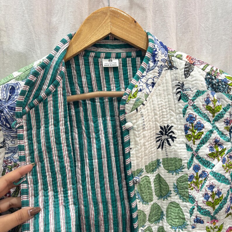 Pu&ograve; includere: Una giacca trapuntata bianca con una fodera a righe verde e bianca. La giacca ha una stampa floreale con fiori blu, verdi e bianchi. La giacca ha una chiusura con bottoni e un colletto. L'etichetta sulla giacca dice "S" e "Made in India".