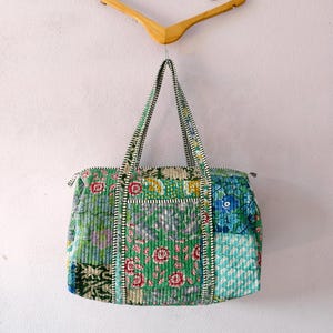 May include: A patchwork tote bag with floral patterns in green, blue, and pink. The bag has black and white striped handles and is suspended from a wooden hanger. The bag's design is a mix of different fabrics.