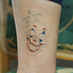 Może przedstawiać: Kolorowy wzór tatuażu przedstawiający czaszkę z koroną. Czaszka ma czerwone usta i akcenty, niebieskie detale oczu i niebieską koronę. Wzór znajduje się na jasnym odcieniu skóry.