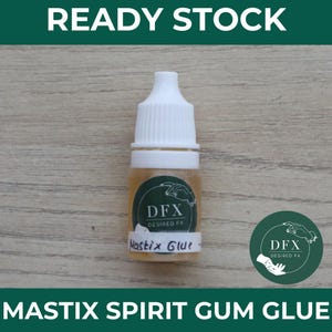 Può includere: Una piccola bottiglia di colla Mastix Spirit Gum con un tappo bianco e un'etichetta che recita "DFX Desired FX". La bottiglia contiene un liquido ambrato chiaro. Sono visibili anche i testi "Ready Stock" e "Mastix Spirit Gum Glue".