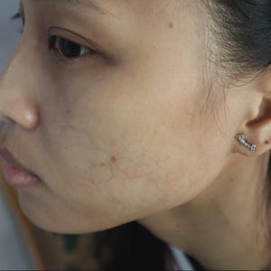 Op de afbeelding: Een zijaanzicht van een gezicht, met de nadruk op de huidtextuur en een kleine, sprankelende oorbel. De huid toont zichtbare aderen en kleine onvolkomenheden. De oorbel is een klein, gebogen ontwerp met meerdere kleine steentjes.