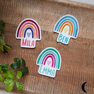 Könnte beinhalten: Drei gestickte Regenbogen-Patches mit Namen. Jeder Patch zeigt ein Regenbogen-Design mit einem Herz und einem Namen: Mila, Ben und Emma. Die Patches liegen auf einer Holzoberfläche.