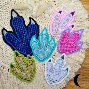 Könnte beinhalten: Eine Sammlung von sechs farbenfrohen Dinosaurier-Fußabdruck-Patches. Die Patches weisen eine Vielzahl von Farben auf, darunter Blau, Grün, Rosa und Lila, mit glitzernden Akzenten. Jeder Patch hat eine schwarze Umrandung und besteht aus Stoff.