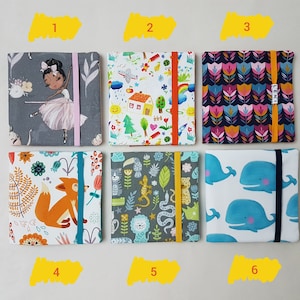 Könnte beinhalten: Sechs stoffbezogene Notizbücher mit elastischen Verschlüssen. Die Notizbücher weisen verschiedene Muster auf, darunter eine Ballerina, ein Haus mit einem Regenbogen, florale Designs, ein Fuchs, Dschungeltiere und Wale. Jedes Notizbuch hat ein unterschiedlich farbiges Gummiband.