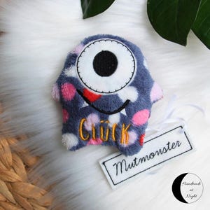 Könnte beinhalten: Ein Plüschmonster-Spielzeug in Blau-, Rosa- und Weißtönen mit einem großen Auge und gestickten Details. Das Wort "Glück" ist mit goldfarbenem Faden gestickt. Ein Etikett lautet "Mutmonster" und "Handmade at Night".