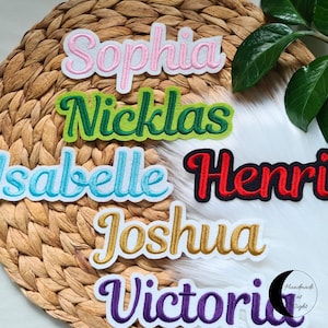 Könnte beinhalten: Gestickte Namensaufnäher in verschiedenen Farben, darunter Rosa, Grün, Blau, Rot, Gold und Lila. Namen sind Sophia, Nicklas, Isabelle, Henri, Joshua und Victoria. Die Aufnäher befinden sich auf einem gewebten Hintergrund.