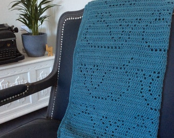 May include: Teal-colored crochet blanket draped over a dark blue armchair. The blanket features a textured pattern with small, evenly spaced holes. A potted plant and a vintage typewriter are visible in the background, adding a touch of vintage charm.