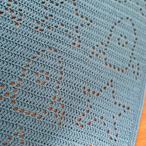 May include: A close-up of a light blue crocheted blanket with a diamond pattern. The blanket has a delicate, openwork design.