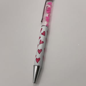 May include: A white pen with a silver cap and a clear barrel filled with pink glitter hearts. The pen has a pink heart pattern on the barrel.
