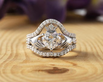 May include: A silver Claddagh ring set with a heart-shaped gemstone, crown, and hands. The ring is encrusted with small, clear gemstones. The ring is set on a wooden surface.