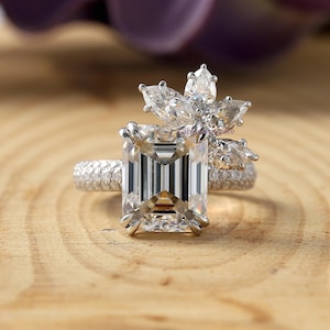 Puede incluir: Primer plano de un anillo de diamantes con una piedra central de talla esmeralda. El anillo presenta un diseño floral con diamantes más pequeños y una banda pavé. El anillo está sobre una superficie de madera.