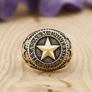 Peut inclure: Une bague dorée avec l'inscription "TEXAS A & M UNIVERSITY" et l'année "2025" gravées. La bague présente une étoile à cinq branches proéminente au centre.