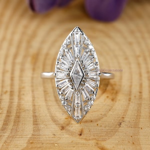 May include: A silver ring with a large, marquise-shaped face. The face is encrusted with numerous small, clear gemstones arranged in a sunburst pattern around a central diamond-shaped stone. The ring is set against a wooden surface.