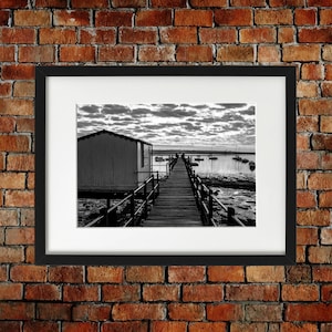 May include: A black and white photograph of a wooden jetty leading to a small building on the water. The jetty is surrounded by railings and there are boats in the distance.