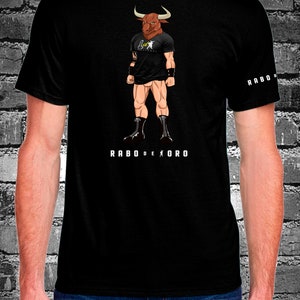 May include: Black t-shirt with a graphic of a bull-headed man wearing a black t-shirt with the text "Rabo de Oro" printed on the front. The text "Rabo de Oro" is also printed on the right sleeve of the shirt.
