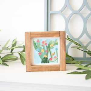 May include: A framed painting of a cactus with pink, yellow, and blue flowers. The cactus is green and has a brown pot. The painting is in a wooden frame.