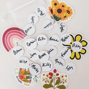 Puede incluir: Una colecci&oacute;n de pegatinas y etiquetas sobre una superficie blanca. Las pegatinas incluyen un arco&iacute;ris, girasoles, un perezoso y un dise&ntilde;o floral. Etiquetas circulares blancas con nombres manuscritos.