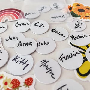 Puede incluir: Una colecci&oacute;n de etiquetas redondas blancas con nombres escritos a mano en tinta negra, incluyendo "Kitty", "Monique" y "Toby". Tambi&eacute;n hay pegatinas decorativas de un arco&iacute;ris, girasoles y fresas.