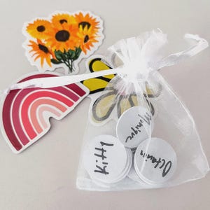 May include: A collection of stickers and small round tokens. Stickers include a rainbow, sunflowers, and a yellow and black butterfly. The tokens are white with black handwritten text and are inside a small white mesh bag.