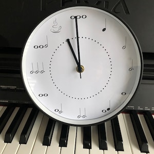 Könnte beinhalten: Eine schwarz-weiße Uhr mit Musiknoten und einem Kaffeetassen-Design. Die Uhrzeiger sind auf 12 Uhr eingestellt.