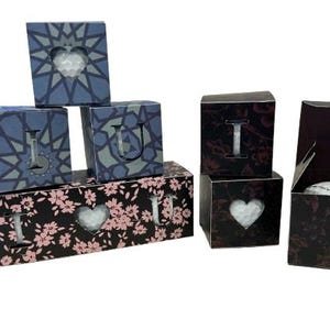 Könnte beinhalten: Anordnung dekorativer Boxen mit Herz- und Buchstabenausschnitten. Einige Boxen sind blau mit geometrischen Mustern, andere schwarz mit floralen Designs. Eine Box ist geöffnet und zeigt ein weißes Objekt.