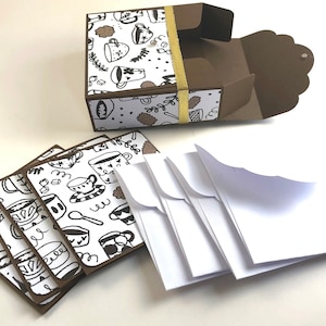 May include: A set of six blank note cards with brown borders and a white background featuring a black and white doodle pattern of coffee cups and mugs. The cards are packaged in a brown box with a gold ribbon and a matching pattern. The box is open and the cards are stacked next to it.