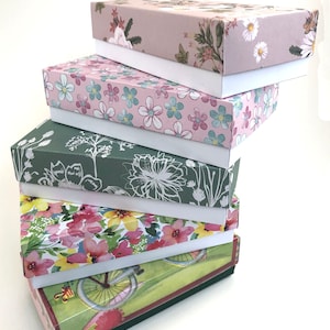 Könnte beinhalten: Ein Stapel von sechs Geschenkboxen mit floralen Mustern und weißen Deckeln. Die Boxen sind pyramidenförmig gestapelt, wobei die größte Box unten und die kleinste Box oben liegt. Die Boxen sind alle mit verschiedenen Blumenmustern verziert, darunter rosa, grüne und weiße Blumen.