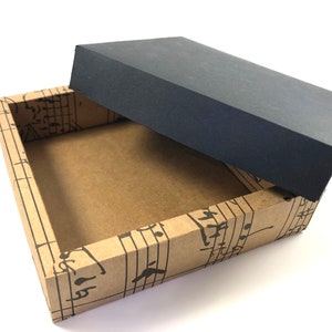 Op de afbeelding: Een bruine kartonnen doos met een zwart deksel. De doos is versierd met een muzieknootpatroon. Het deksel is open en de binnenkant van de doos is leeg.