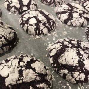 May include: A baking sheet filled with freshly baked chocolate cookies covered in powdered sugar. The cookies have a cracked and textured surface, giving them a unique appearance.