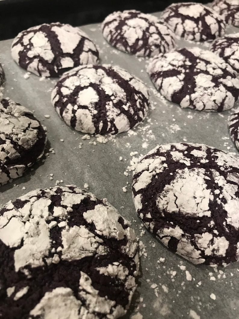 May include: A baking sheet filled with freshly baked chocolate cookies covered in powdered sugar. The cookies have a cracked and textured surface, giving them a unique appearance.