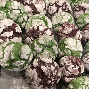 May include: A close-up of a variety of green and brown cookies with a white powdered sugar coating. The cookies are arranged on a white parchment paper-lined baking sheet.