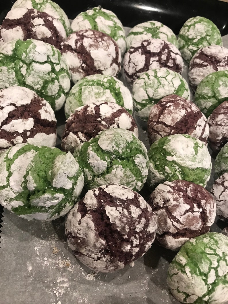May include: A close-up of a variety of green and brown cookies with a white powdered sugar coating. The cookies are arranged on a white parchment paper-lined baking sheet.