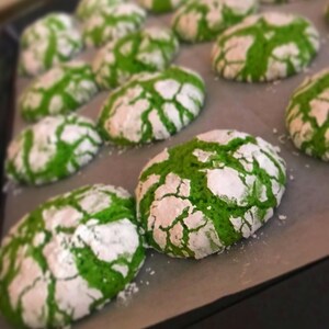May include: A baking sheet lined with parchment paper filled with green and white cookies. The cookies have a cracked and crinkled surface, and are dusted with powdered sugar.