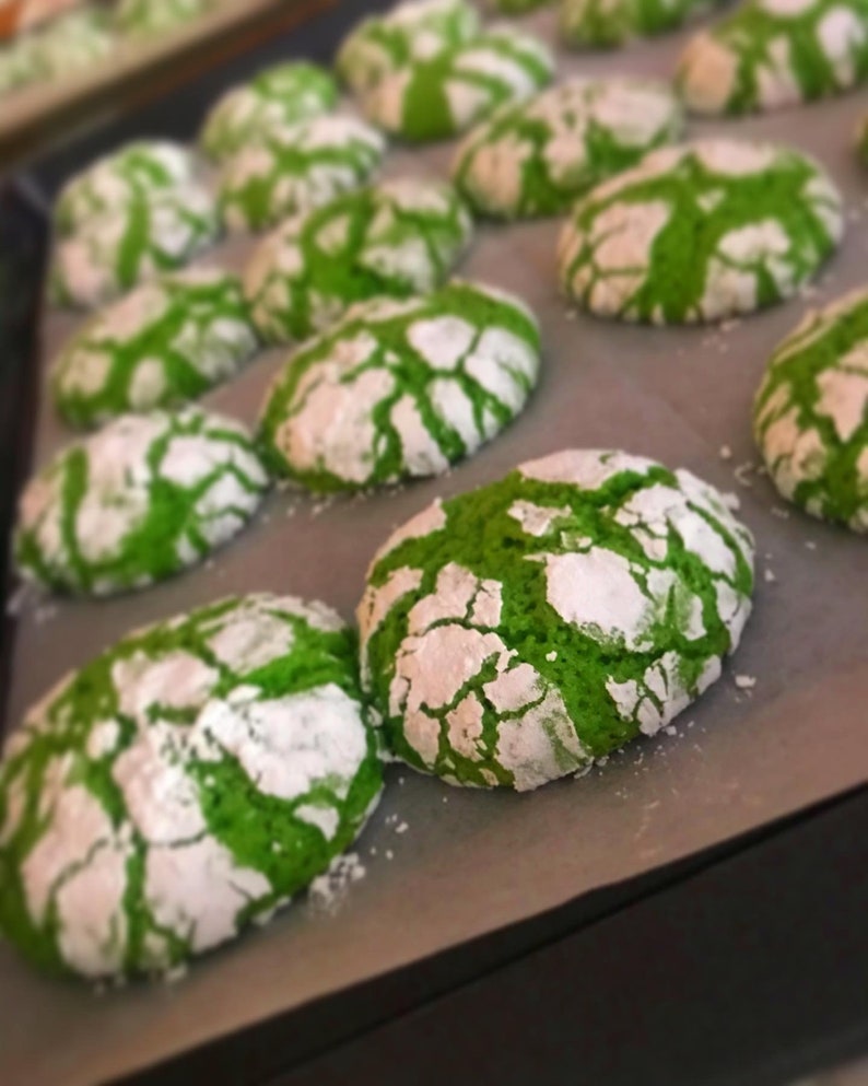 May include: A baking sheet lined with parchment paper filled with green and white cookies. The cookies have a cracked and crinkled surface, and are dusted with powdered sugar.