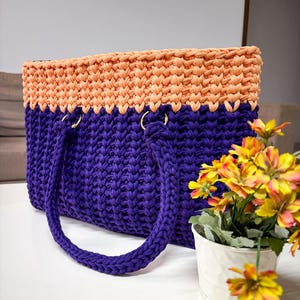 May include: A handmade tote bag in a two-tone design. The top section is peach-colored, while the main body is a deep purple. The bag features two rounded handles and gold-colored rings. The bag is sitting on a white surface.