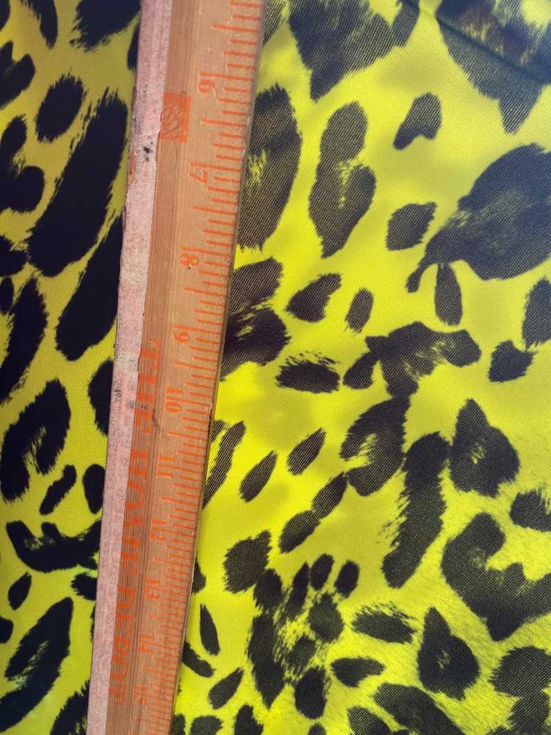 Puede incluir: Primer plano de una tela amarilla con un estampado de leopardo negro. Una regla de madera se coloca sobre la tela para mostrar la escala.