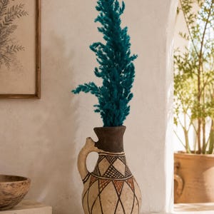 May include: A tall, bulbous ceramic pitcher with a single handle, featuring a cream-colored base decorated with brown and black geometric diamond patterns. The neck is dark brown, and it holds a tall, vibrant teal-colored pampas grass plume.