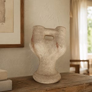 May include: A beige, textured ceramic vase with a sculptural, organic form featuring two rounded top openings connected by a central bridge. The surface has a matte, stone-like finish with subtle reddish-brown color variations on the sides.