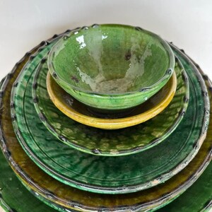 May include: A stack of five glazed ceramic dishes consisting of four round plates and one small bowl. The pieces feature a glossy finish in shades of deep forest green and mustard yellow, with dark, scalloped, and textured edges on each item.