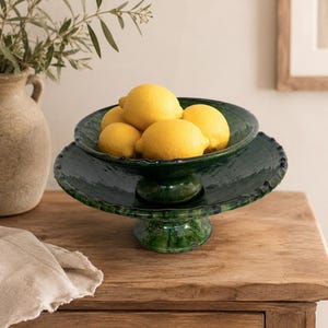 Peut inclure: Un bol sur pied en céramique émaillée vert foncé avec un bord festonné, rempli de quatre citrons jaune vif. Le bol présente une finition brillante et irrégulière et repose sur une base évasée robuste, affichant une teinte émeraude profonde.