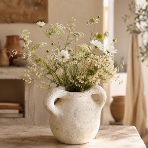 Pode incluir: Um vaso de cerâmica texturizada na cor creme com duas alças laterais proeminentes, preenchido com um buquê de flores silvestres brancas e verde-pálido, incluindo gipsófila delicada, cosmos e cenoura selvagem, num arranjo natural e arejado.