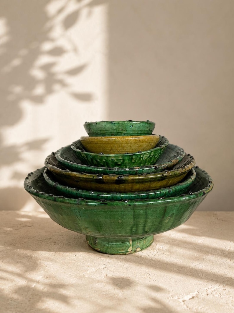 May include: A stack of six nested ceramic bowls with a glossy glaze in shades of emerald green and olive yellow. Each bowl features a wide, shallow basin, a pedestal base, and a decorative scalloped rim with small, raised bumps along the edge.