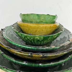 May include: A stack of five glazed ceramic bowls and plates in varying shades of deep forest green and mustard yellow. Each piece features a scalloped, textured rim with a glossy finish, creating a layered appearance of nested circular vessels.
