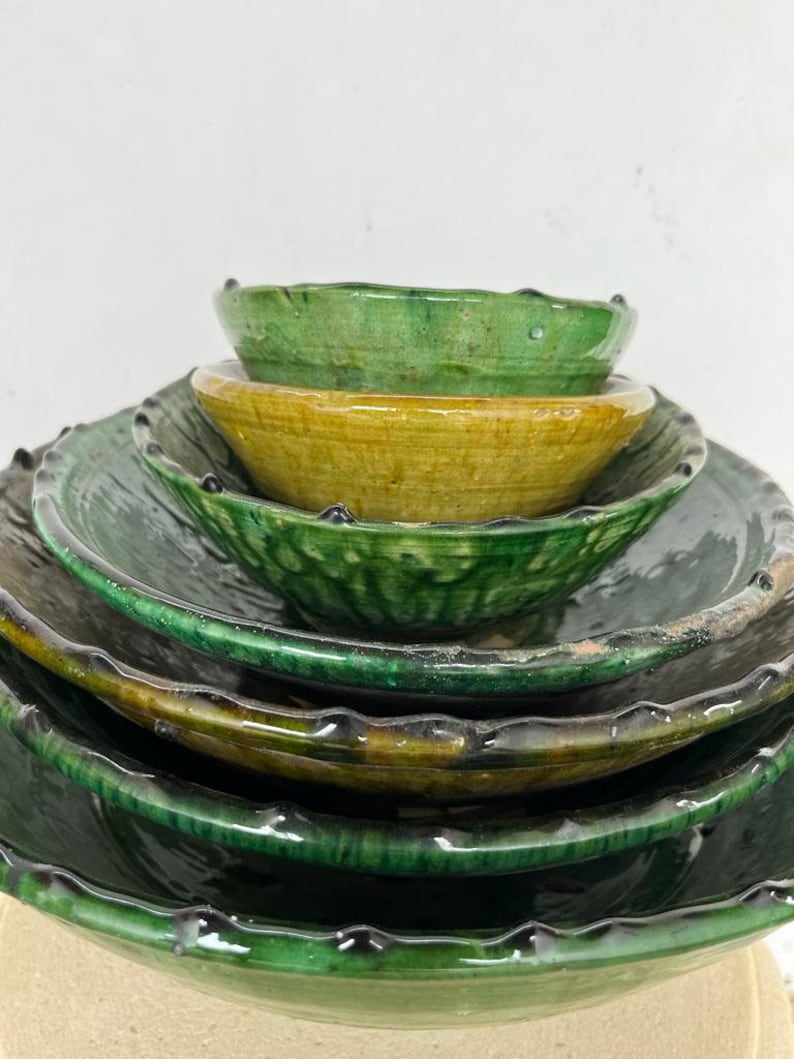 May include: A stack of five glazed ceramic bowls and plates in varying shades of deep forest green and mustard yellow. Each piece features a scalloped, textured rim with a glossy finish, creating a layered appearance of nested circular vessels.