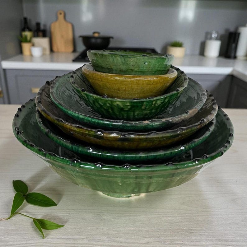 May include: A set of five nested ceramic bowls with a glossy finish. The bowls feature a scalloped edge design and are colored in varying shades of deep forest green and a single mustard yellow bowl positioned near the top of the stack.