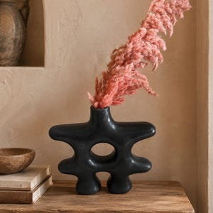 Peut inclure: Un vase en céramique noir mat avec une forme abstraite à plusieurs lobes et une découpe circulaire centrale. Le vase contient une grande tige plumeuse d'herbe de la pampa rose séchée, créant un contraste saisissant entre le récipient sombre et lisse et l'élément botanique texturé.