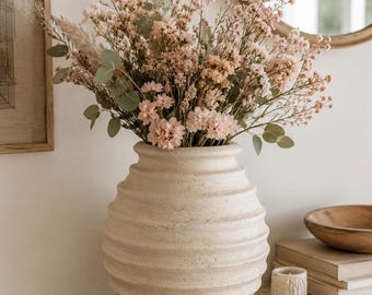 Pode incluir: Um vaso de cerâmica bege, texturizado e arredondado, com sulcos horizontais, preenchido com um grande arranjo de flores secas cor-de-rosa, pequenas flores brancas e folhas de eucalipto verdes, criando um arranjo floral de tons neutros.